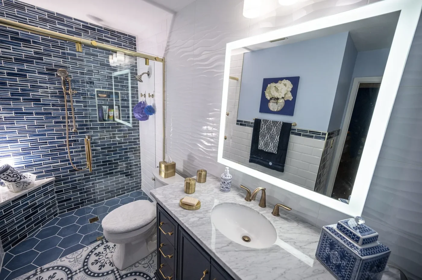 Bathroom after remodeling in St. Petersburg by CMK with blue glass mosaic shower, navy vanity, Carrara marble top, and lighted mirror