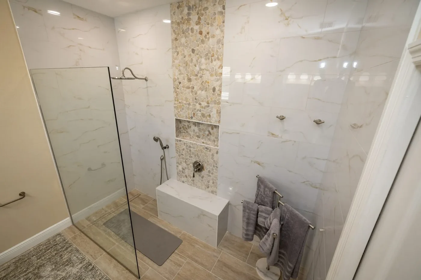 Curbless walk in shower after remodeling in Tampa by CMK with marble look porcelain, pebble accent column with niche, tiled bench, linear drain, and rain shower head