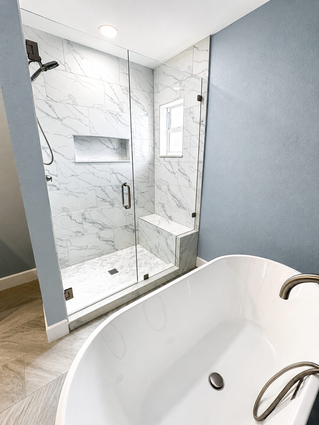 Walk in shower with marble look wall tile, tiled bench, and herringbone mosaic floor beside a freestanding tub in a Clearwater master bathroom remodel