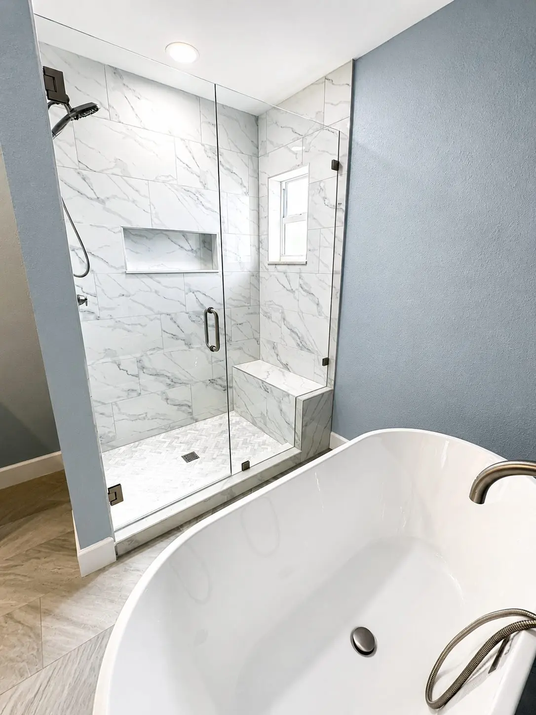 Walk in shower with marble look wall tile, tiled bench, and herringbone mosaic floor beside a freestanding tub in a Clearwater master bathroom remodel