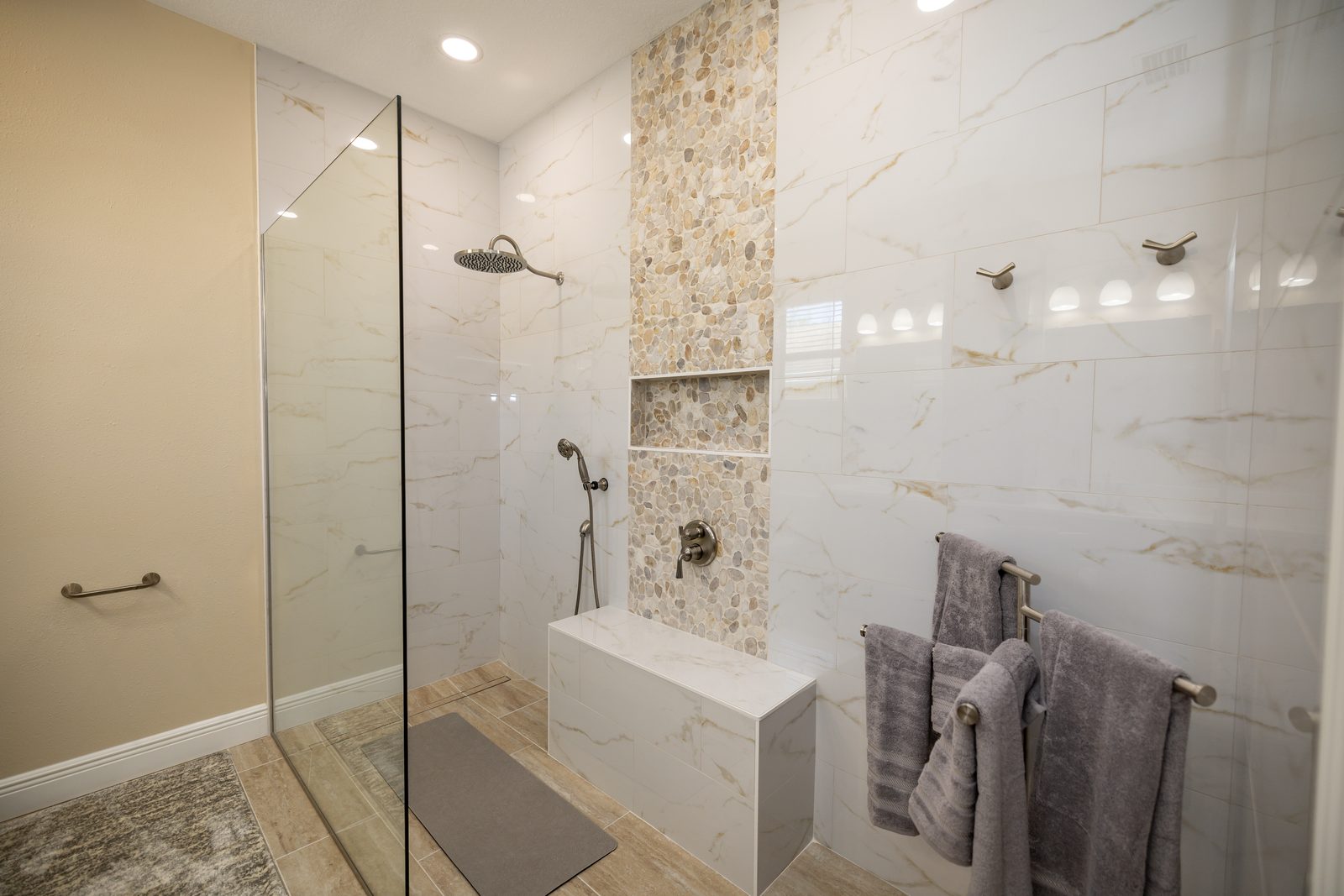 Curbless walk in shower with gold veined wall tile, vertical pebble mosaic accent column through the niche, tiled bench, and wood look plank floor with linear drain in an Indian Rocks Beach remodel