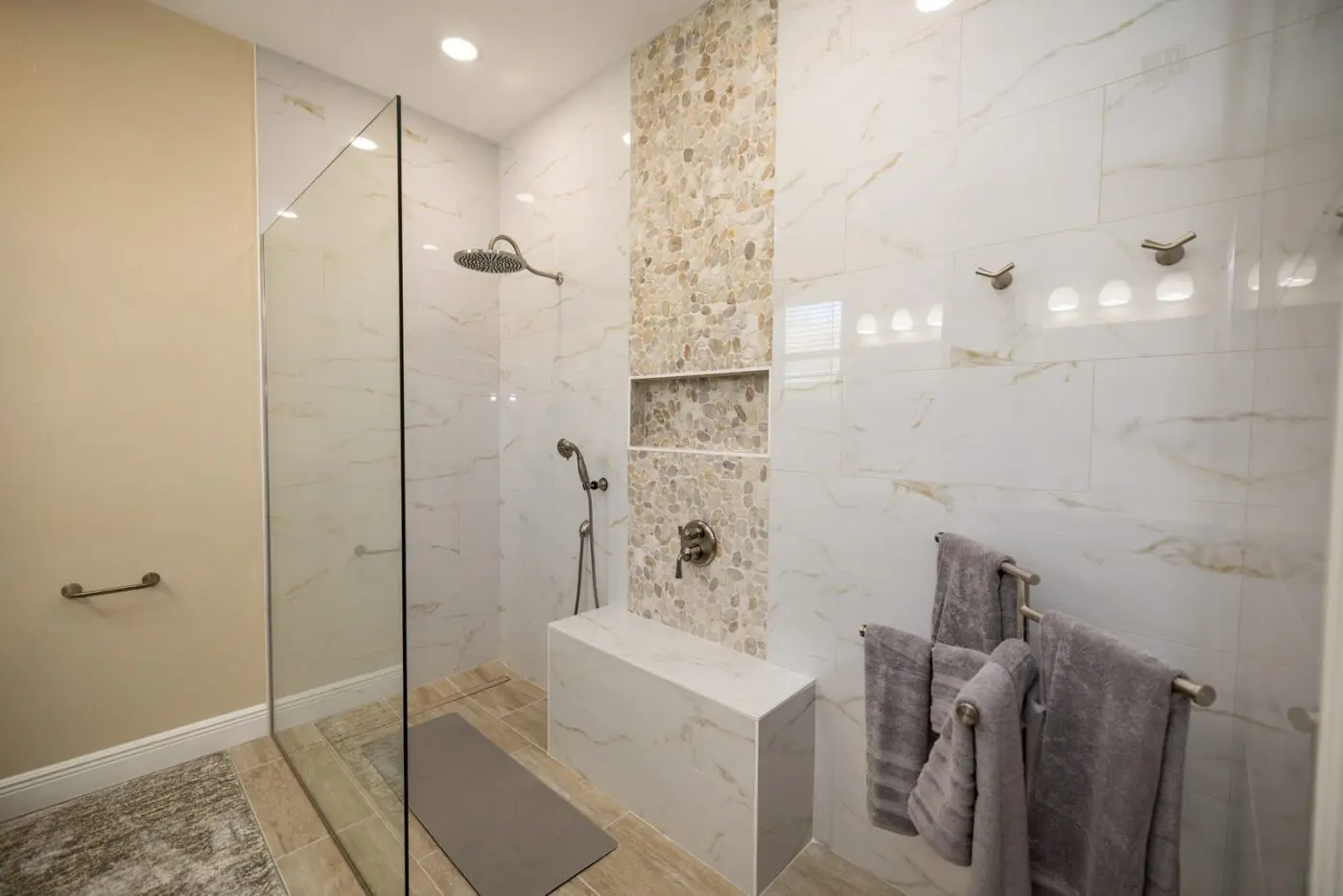 Curbless walk in shower with gold veined wall tile, vertical pebble mosaic accent column through the niche, tiled bench, and wood look plank floor with linear drain in an Indian Rocks Beach remodel