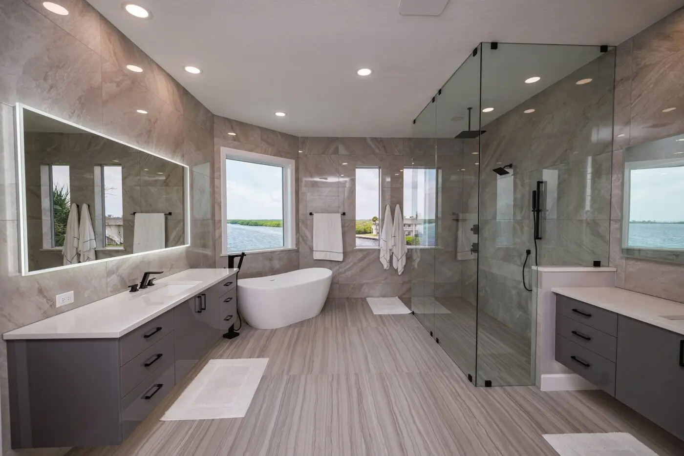 Waterfront master bathroom with stone look tile drenched walls, wood look plank floor tile running into a frameless glass shower, freestanding tub, and floating vanities in a Safety Harbor remodel