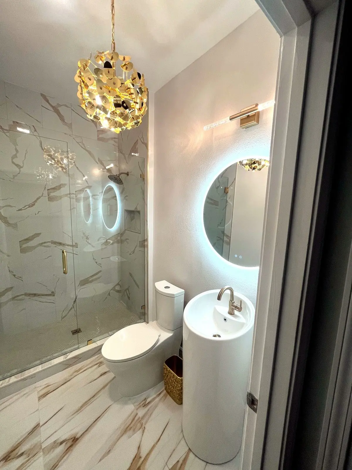Hall bathroom with gold veined marble look tile on shower walls and floor, pedestal sink, and gold light fixtures in a Tarpon Springs remodel