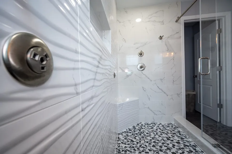 Corner shower bench faced in sculpted wave tile with marble look walls and pebble mosaic floor in a Palm Harbor shower