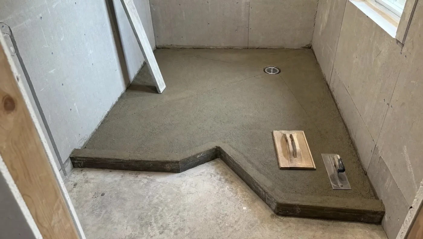 Custom L-shaped mortar bed shower pan sloped to an offset drain, with a wood float and steel trowel resting on the fresh deck mud