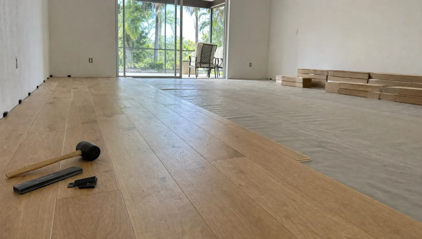 Light oak engineered wood flooring half installed over a moisture-barrier underlayment on a concrete slab after the walls are primed, with flooring boxes, a mallet and spacers, and sliding doors to a screened lanai