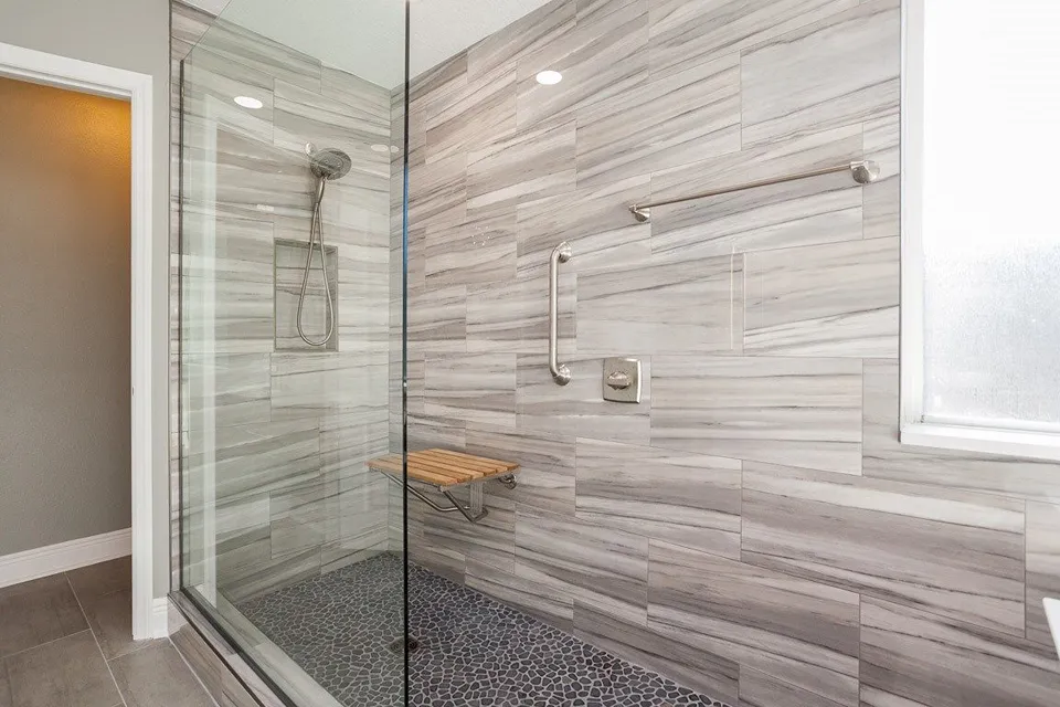 Wall mounted fold down teak shower seat with grab bar on gray wood look tile and pebble floor in a Tarpon Springs shower