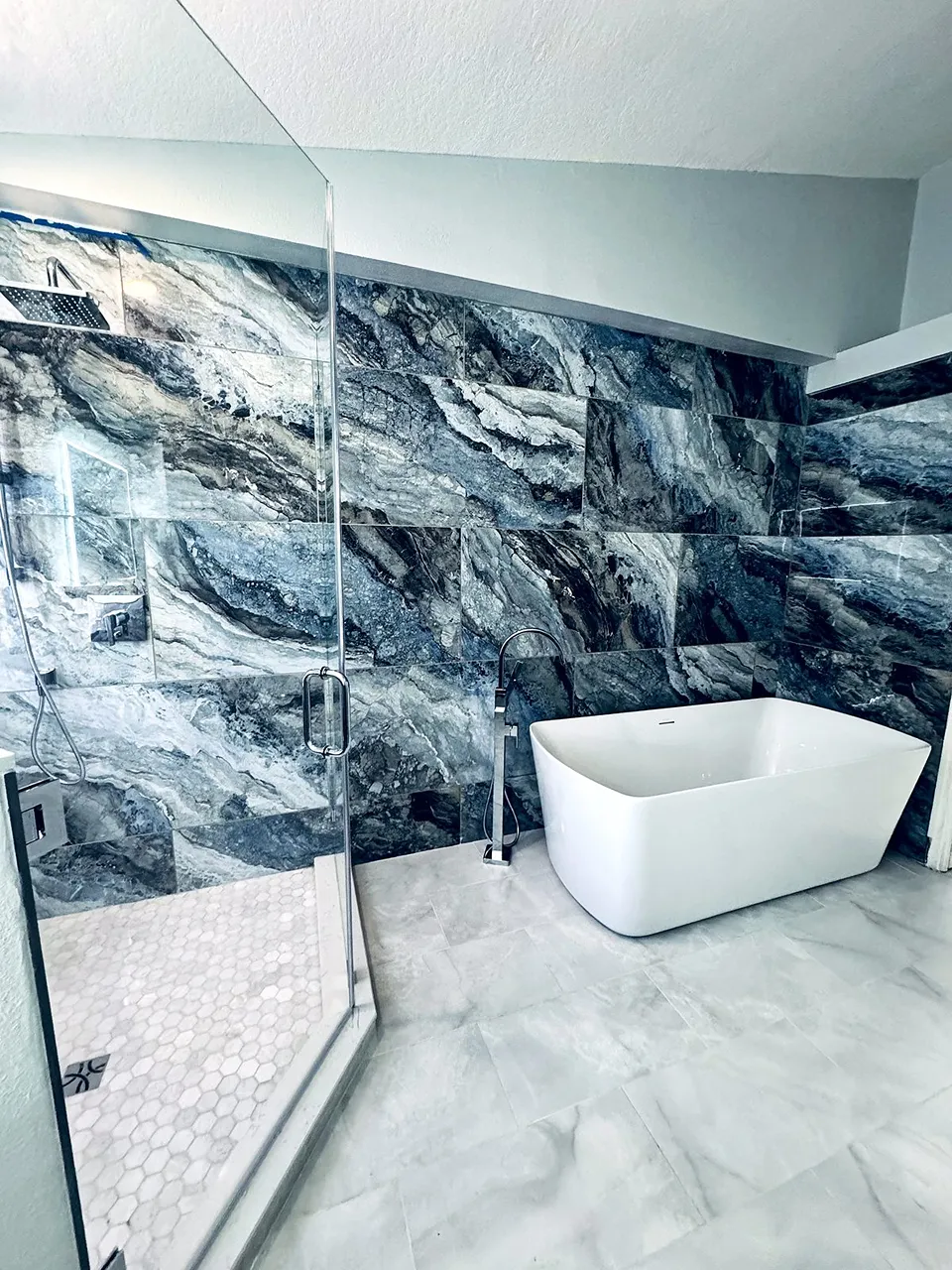 Rectangular freestanding soaking tub against a blue and gray book-matched large-format porcelain statement wall beside a frameless glass shower in a Palm Harbor bathroom
