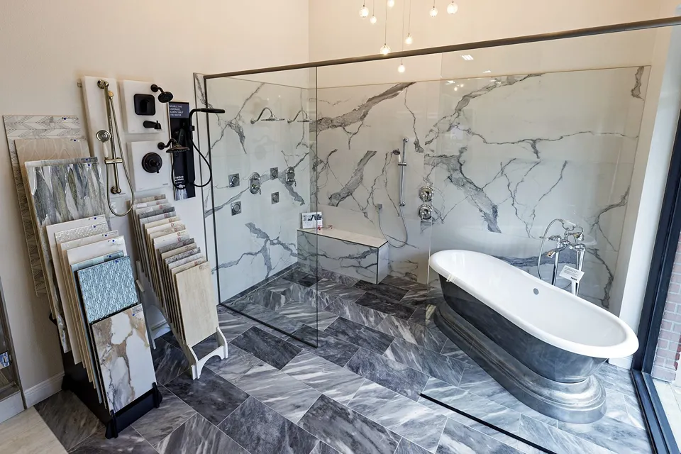 Metallic pedestal freestanding tub with a chrome telephone filler at the edge of a curbless walk-in wet room with marble-look tile and linear drain in the CMK Tampa Design Studio