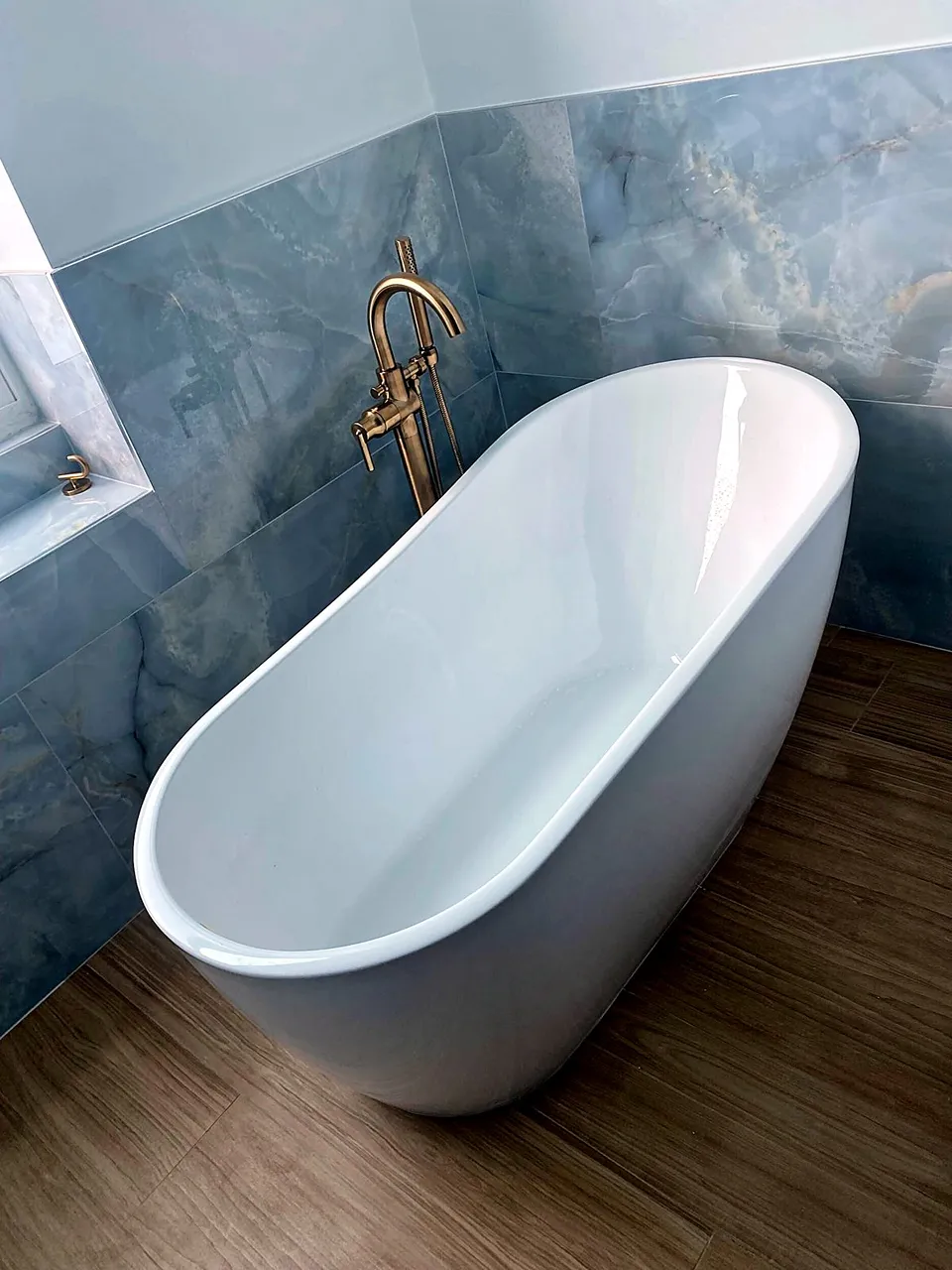 Oval freestanding slipper tub with a champagne bronze floor-mounted filler and hand shower against blue onyx-look large-format porcelain in a Tampa bathroom remodel