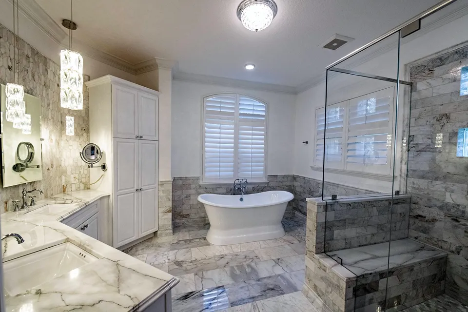 Double-slipper freestanding pedestal tub centered in a tiled alcove under an arched shuttered window in a marble Tampa owner's suite bathroom