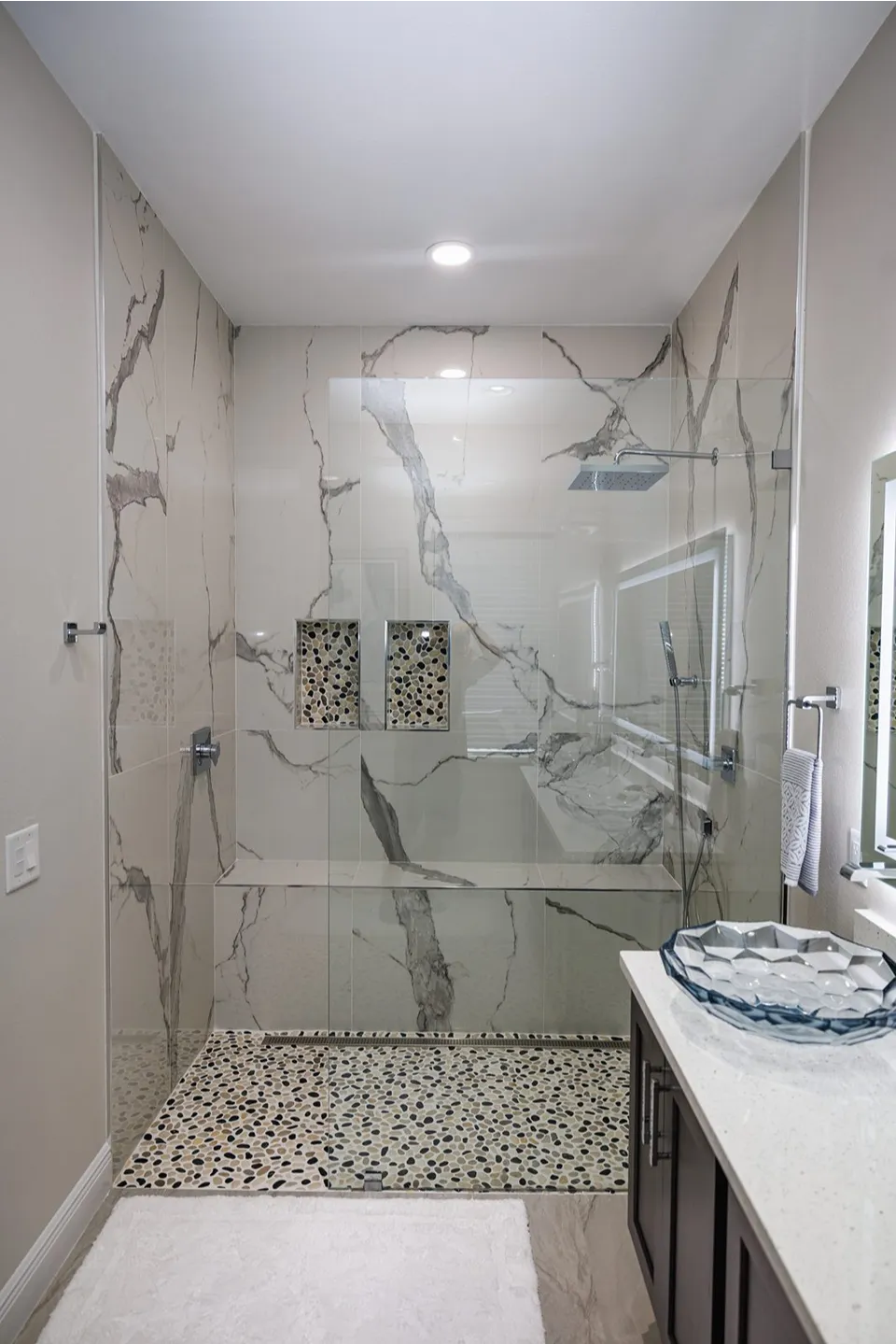 Full width built in shower bench in statuario look porcelain with pebble lined niches and linear drain in a Tampa walk in shower