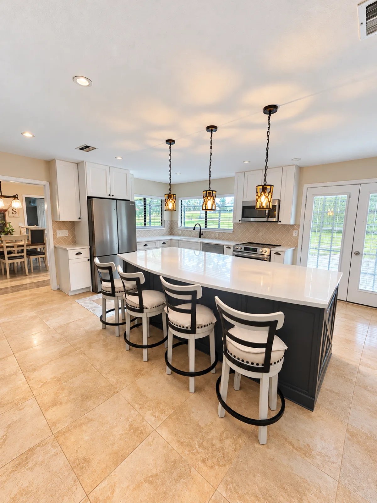 Finished Hudson kitchen seen from the dining side, with the dark island, six white stools, pendant lights, and French doors to the lanai