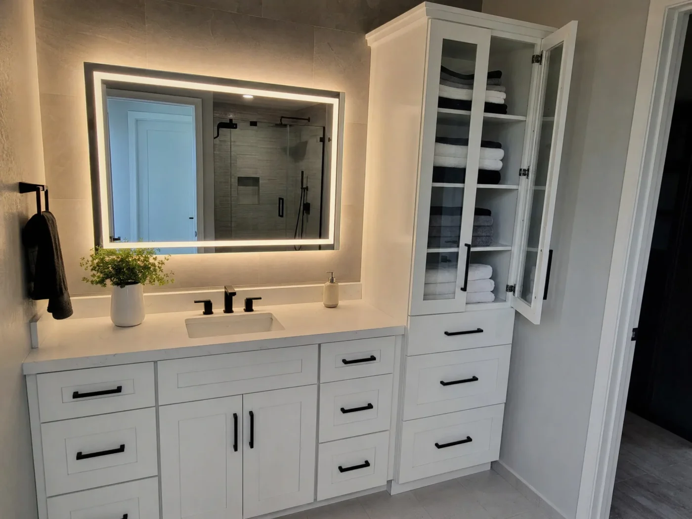 LED backlit mirror with adjacent storage tower above white shaker vanity with matte black hardware in Wesley Chapel FL bathroom by CMK Construction