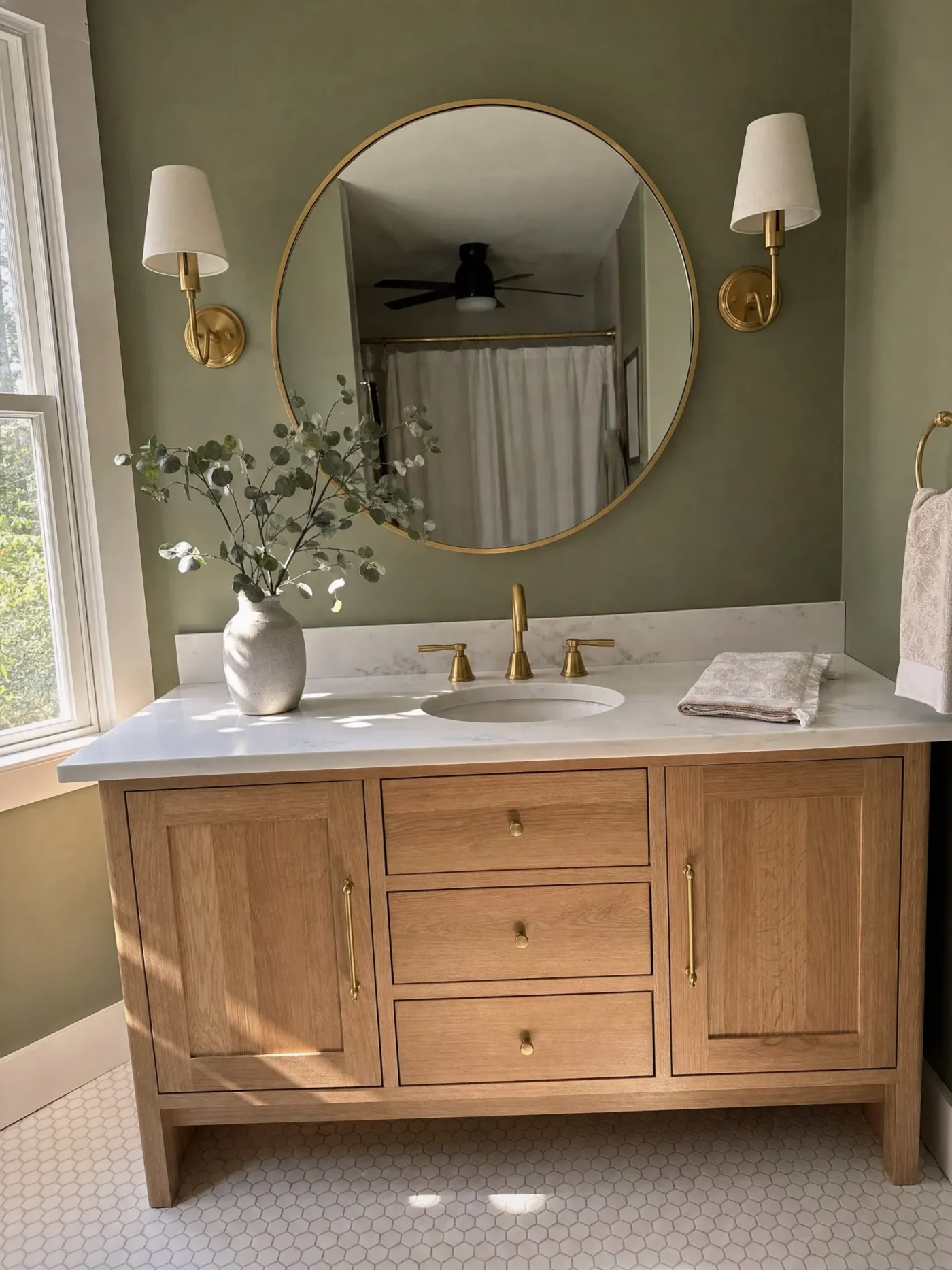 Large round mirror with brass frame flanked by wall sconces above white oak vanity with marble countertop in Palm Harbor FL bathroom by CMK Construction