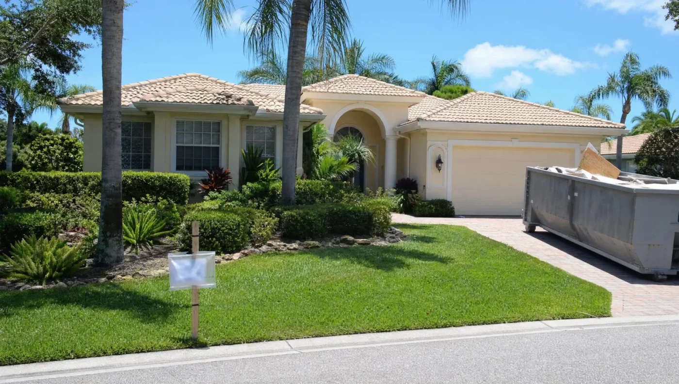 1990s stucco home in Sarasota with a tile roof during an interior remodel, with a building permit box posted by the driveway and a roll-off dumpster