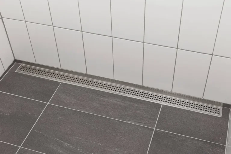 Finished installation of a stainless steel linear shower drain positioned along white tiled shower walls and gray floor tiles.