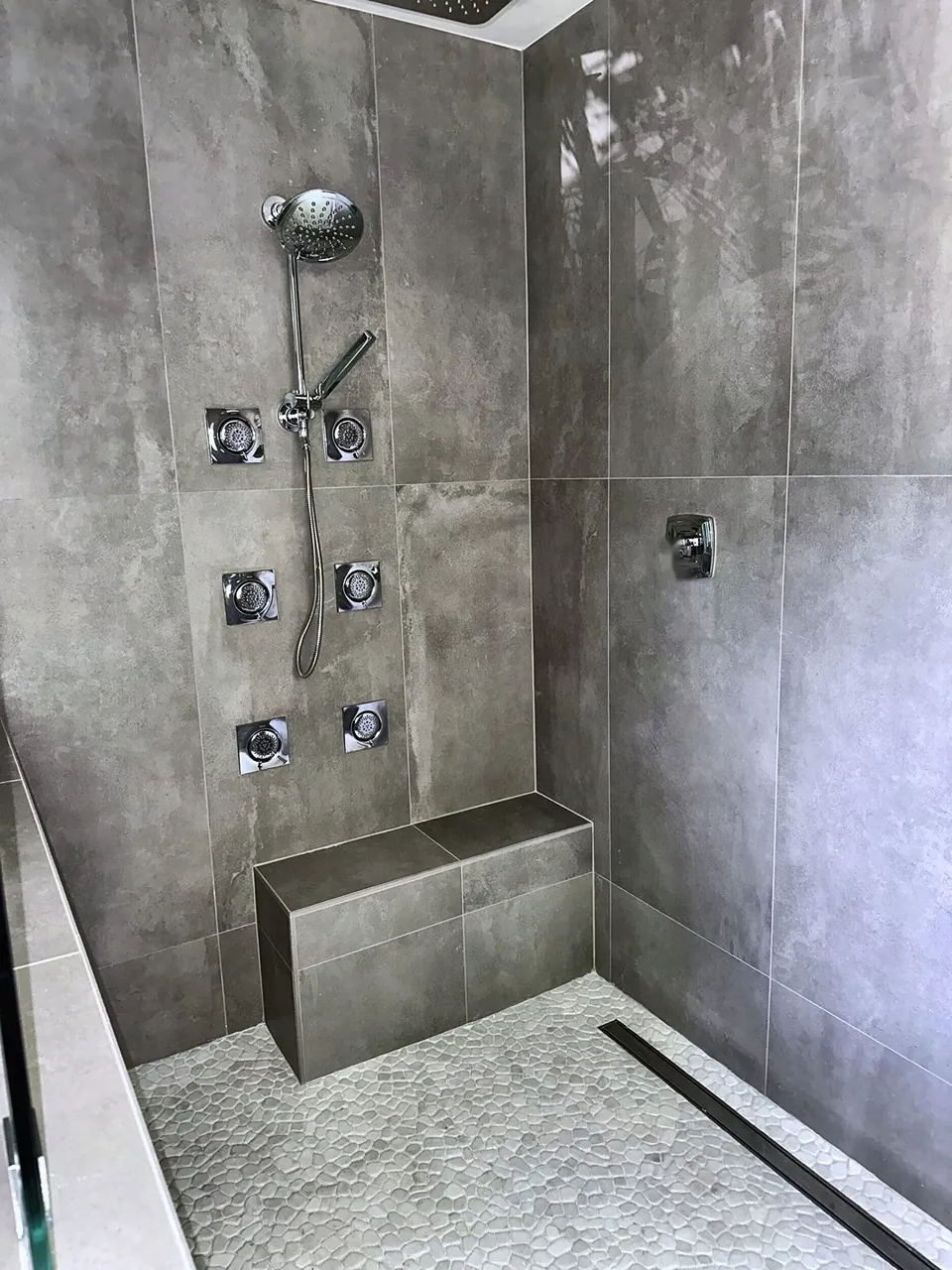 Tiled shower bench beneath body sprays with white pebble stone floor and linear drain in a Safety Harbor wet room shower