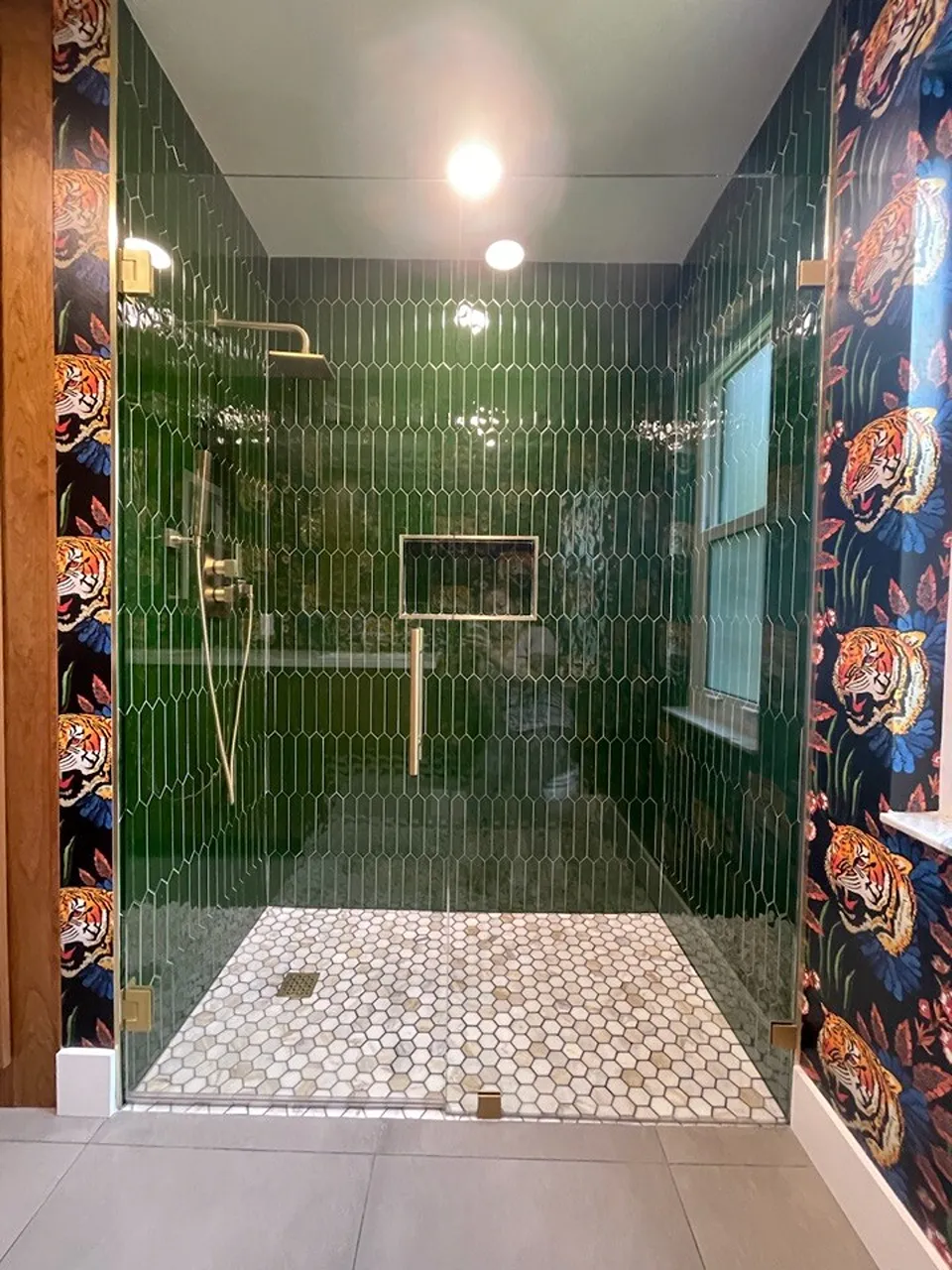 Emerald green glossy elongated picket tile covering the walls of a walk in shower with brushed gold rain head, gold framed niche, and white marble hexagon mosaic floor in an Oldsmar FL bathroom remodel by CMK Construction