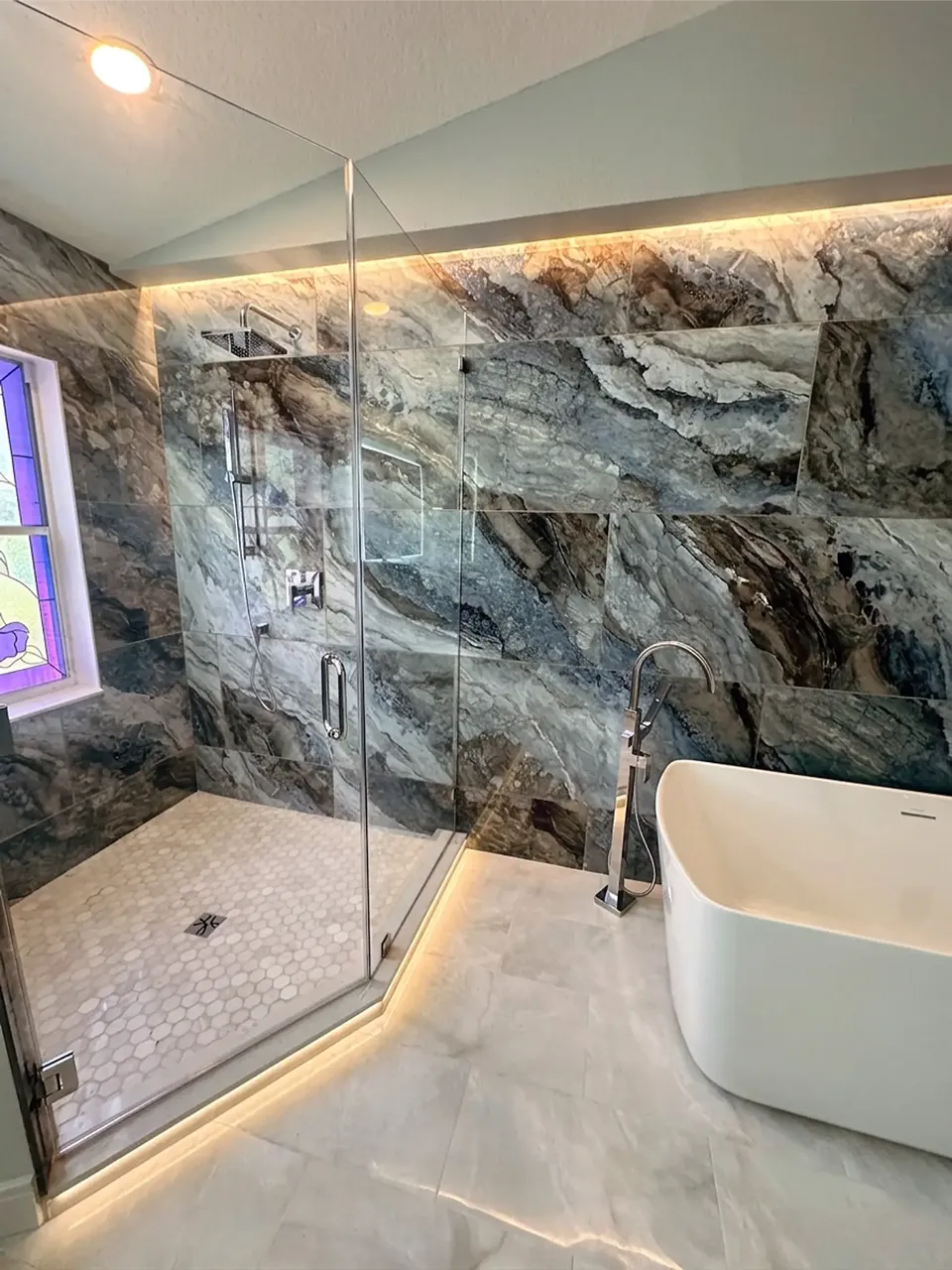Dramatic blue and brown quartzite look large format shower tile continuing onto the tub wall with LED cove lighting, white hexagon mosaic shower floor, and a freestanding tub in a Palm Harbor FL primary bathroom remodel by CMK Construction