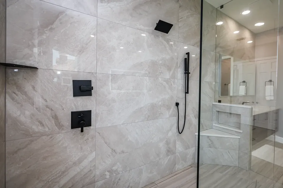 Polished large format greige marble look shower tile with a matte black square rain head, black slide bar handheld, black corner shelf, and a built in corner bench with a stone look slab top in a Siesta Key FL bathroom remodel by CMK Construction