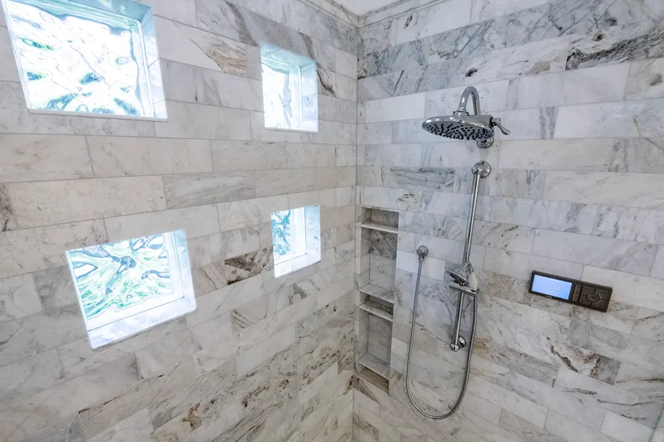 White and gray marble subway tile set in a brick pattern with four lit art glass block windows, a recessed corner shelving column, chrome rain head, and a digital shower control in a Tampa FL bathroom remodel by CMK Construction