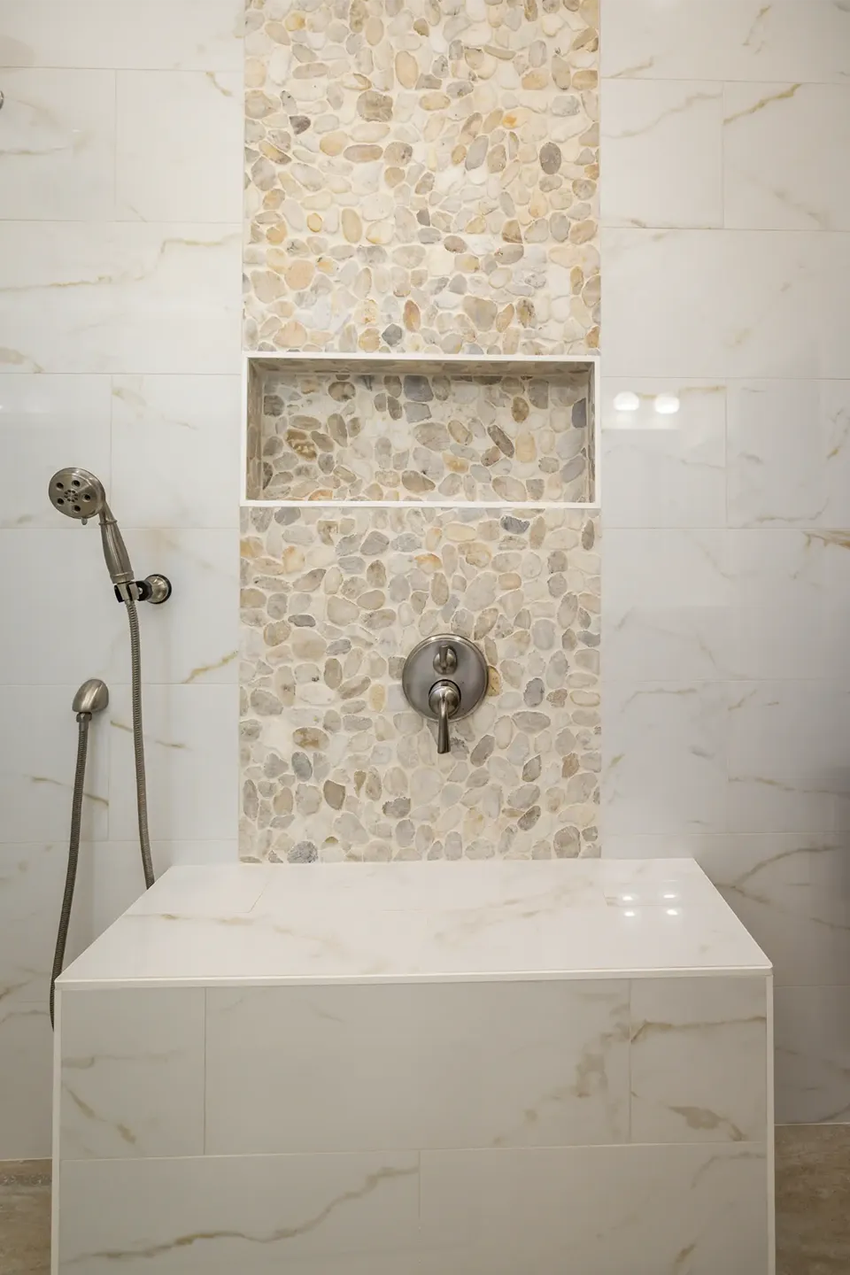 Vertical pebble mosaic accent column running floor to ceiling through a recessed niche and valve wall between gold veined marble look shower tile above a full width tiled bench in a Tarpon Springs FL bathroom remodel by CMK Construction