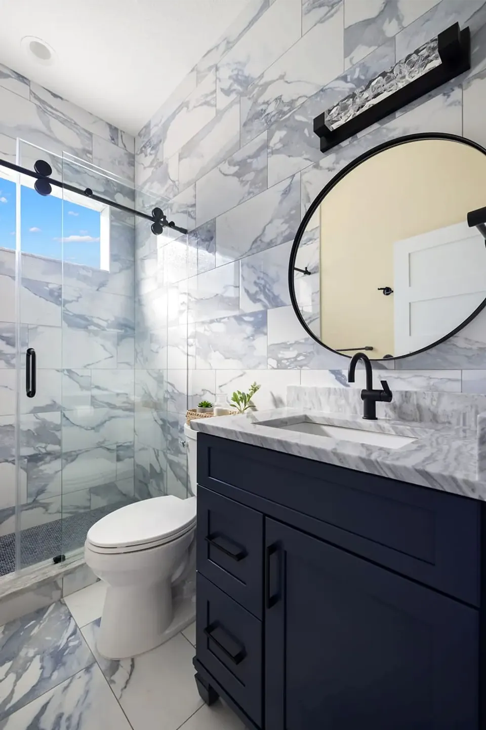 Arabescato marble look large format tile wrapping the shower and vanity wall with a matte black barn style sliding glass door, navy shaker vanity, and round black mirror in a Trinity FL bathroom remodel by CMK Construction
