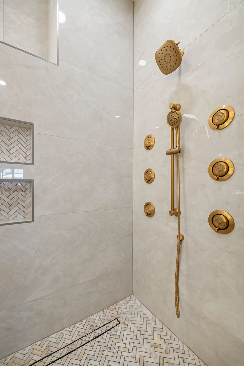 Cream marble look large format shower tile with brushed gold body sprays, stacked herringbone mosaic niches, and a herringbone mosaic floor with linear drain in a Trinity FL primary bathroom remodel by CMK Construction