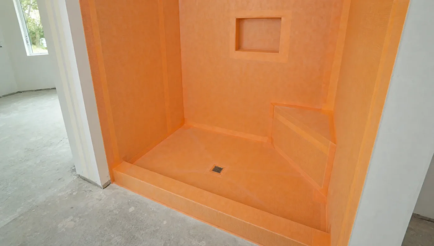 Walk-in shower covered in an orange bonded waterproofing membrane over the walls, niche, corner bench, sloped floor and curb before tile is installed