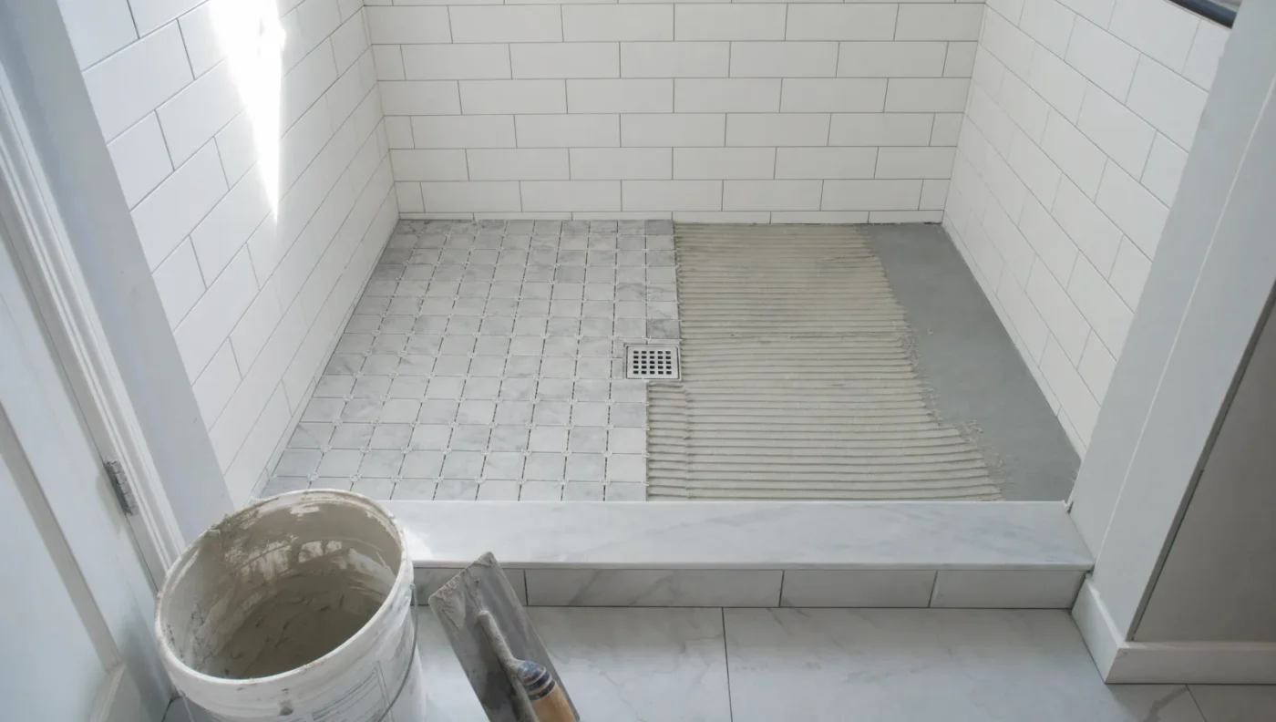 Tile-ready shower pan half covered in gray mosaic tile with combed thinset on the other half, a square center drain, and white subway tile walls