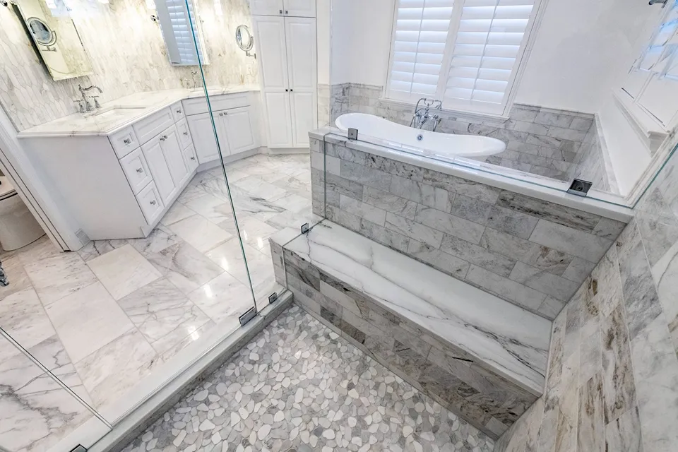 Stone slab topped shower bench beside a freestanding tub half wall behind frameless glass with pebble floor in a Tampa walk in shower