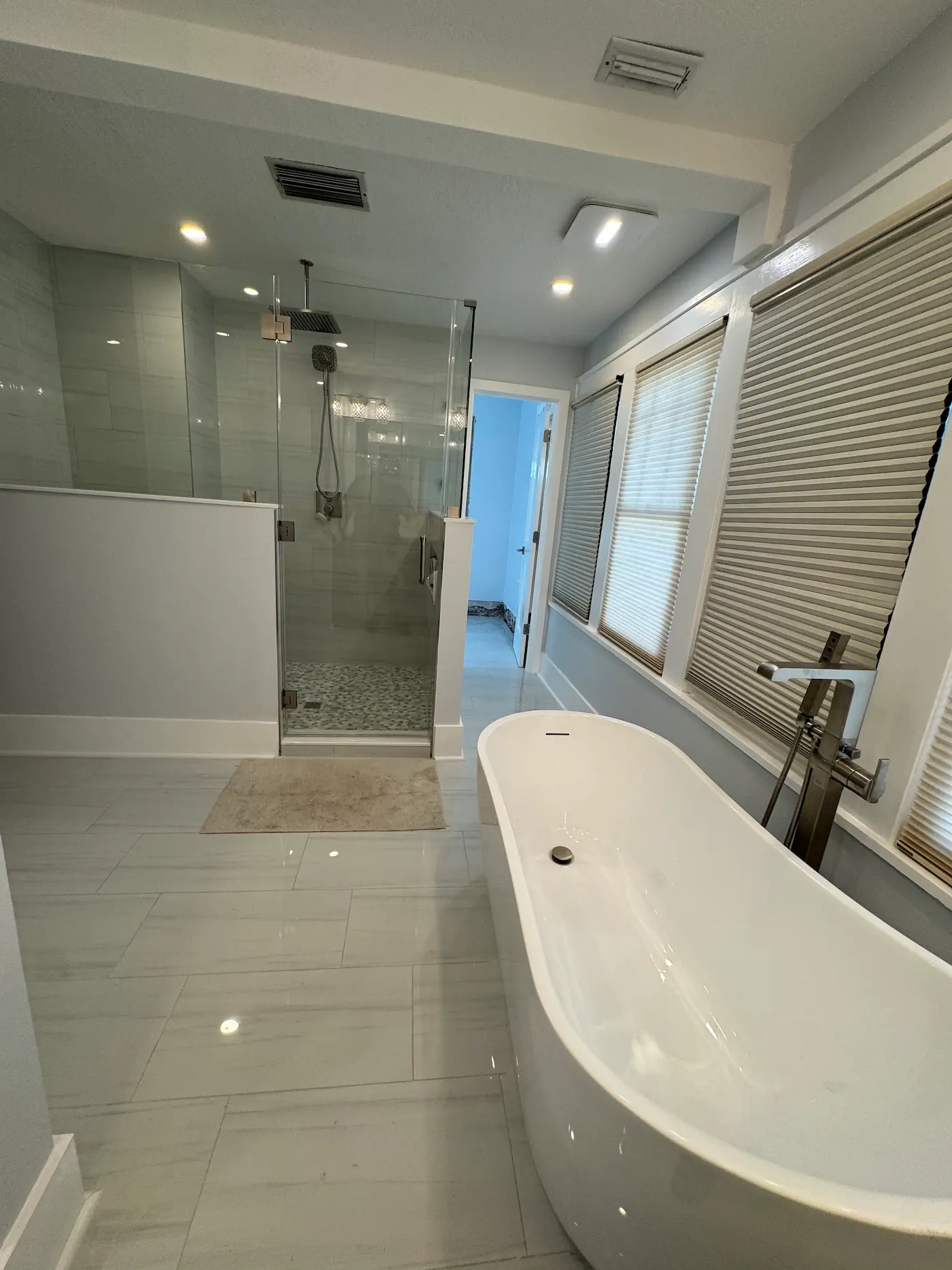 Primary bathroom with a glass walk-in shower set between two white pony walls, a ceiling-mounted rain head plus a handheld on a slide bar, a pebble shower floor, and a freestanding tub in front