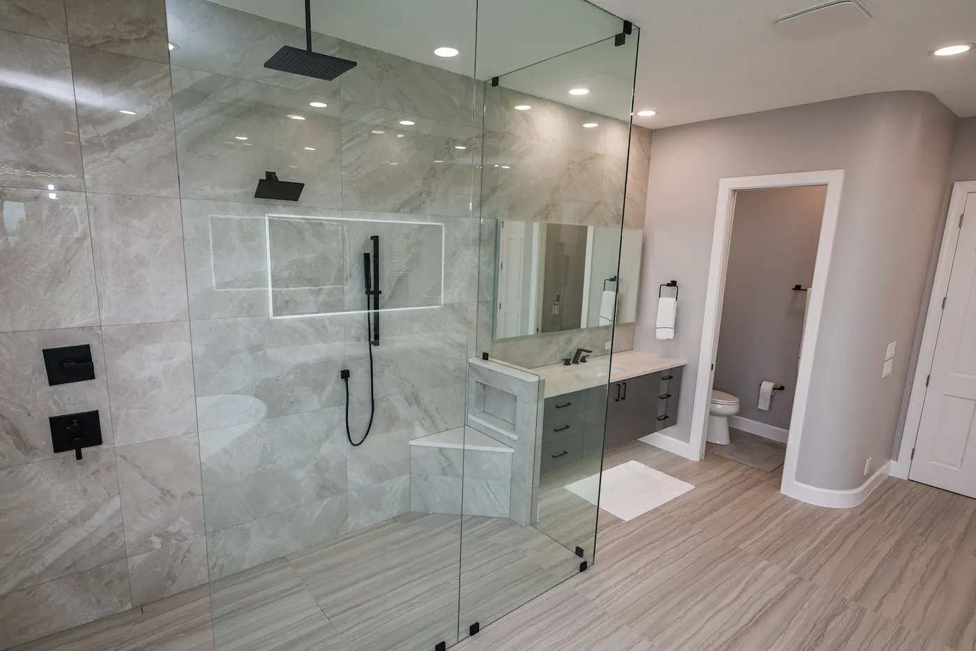 Curbless walk-in shower with fixed frameless glass panels, gray large-format tile, matte black rain head and handheld, lit niche, and a corner bench next to a floating vanity