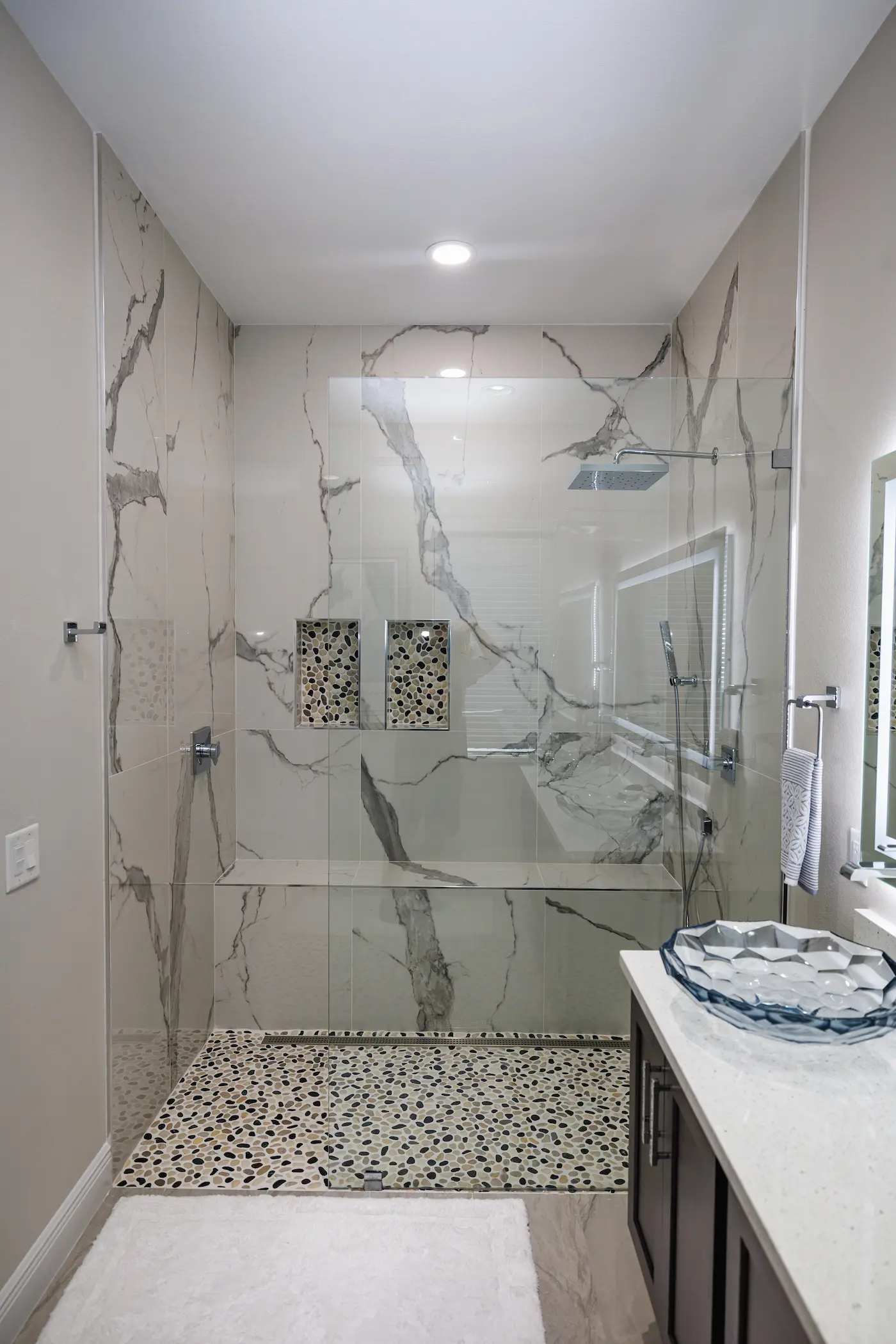 Doorless walk-in shower with one fixed glass panel, white marble-look porcelain walls, a linear drain along the back wall, a pebble tile floor, and two pebble-lined niches