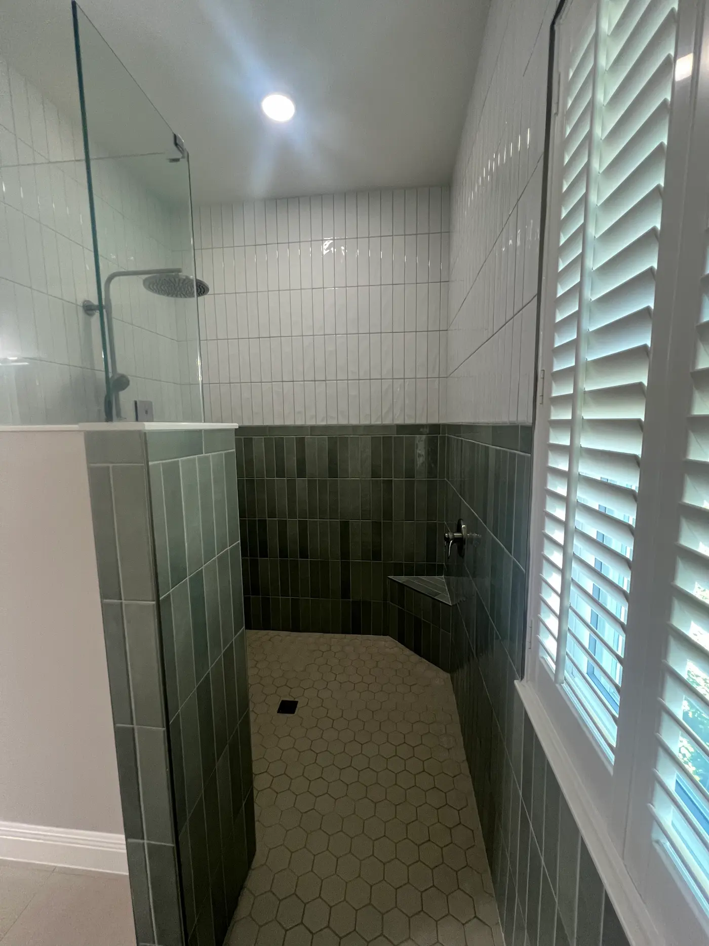 Doorless walk-in shower entered past a tiled half wall with a glass panel above, green vertical stacked tile below white vertical stacked tile, a hexagon floor, a corner bench, and a shuttered window