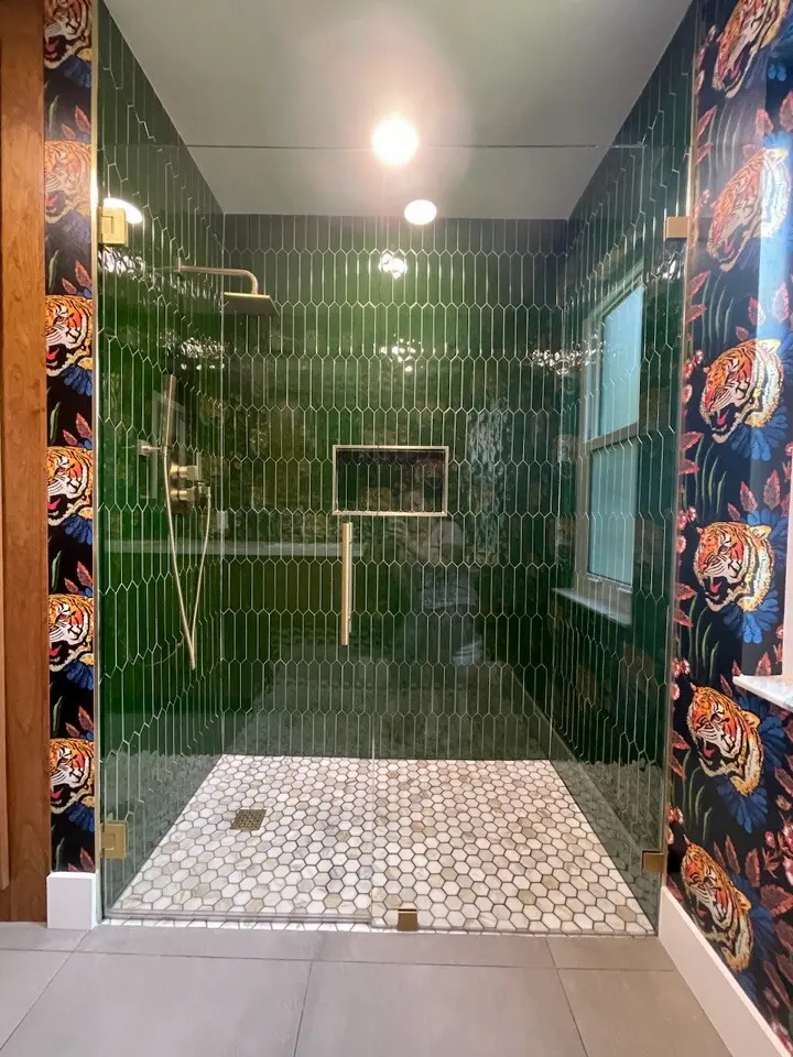Walk-in shower with glossy emerald green elongated hexagon tile, brass fixtures and glass hardware, a white hexagon mosaic floor, a recessed niche, a window, and tiger print wallpaper outside the glass