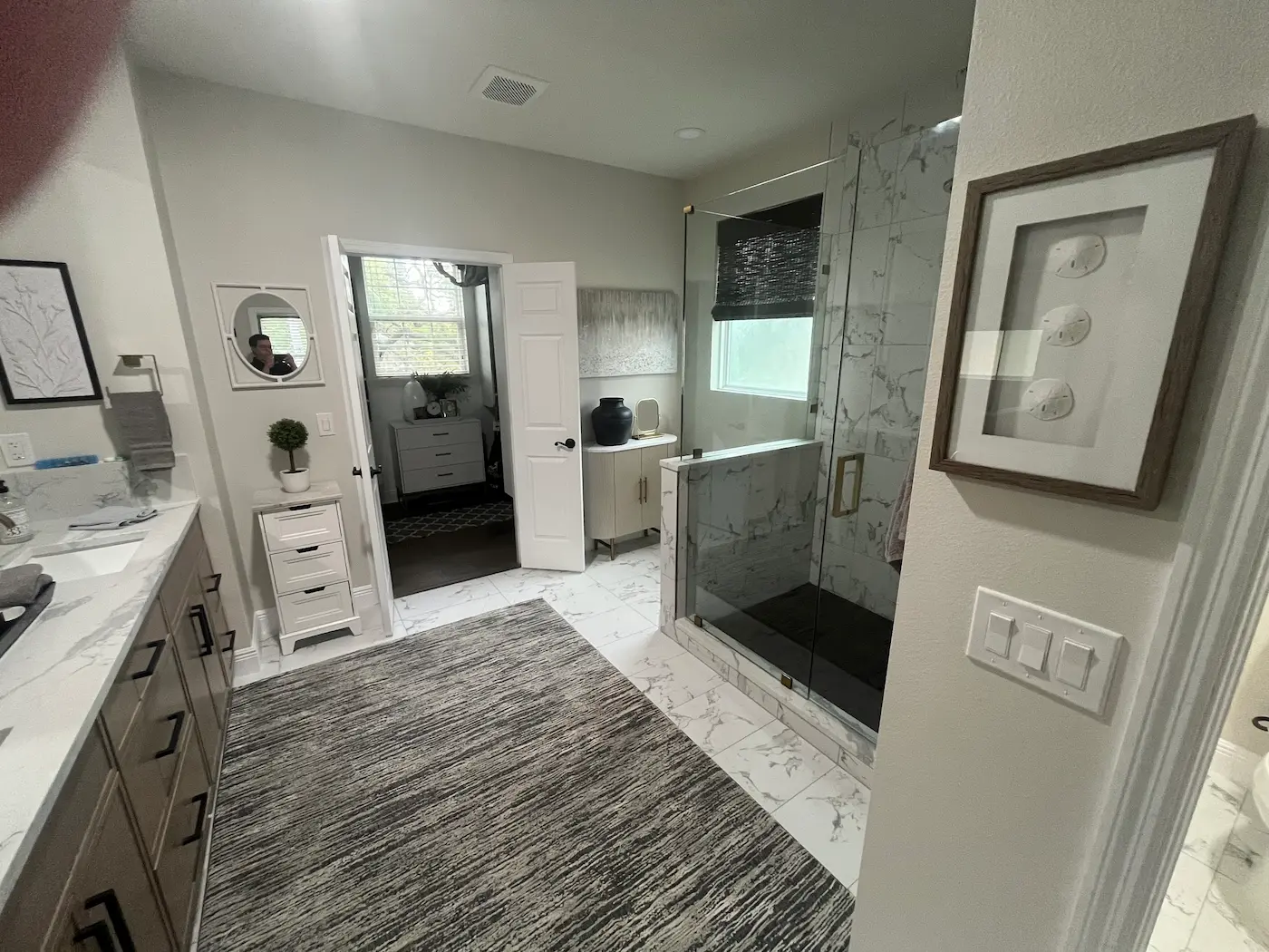 Primary bathroom with a walk-in shower behind a white marble-look tiled half wall topped with glass, brass door handle and clips, a window inside the shower, and a dark shower floor
