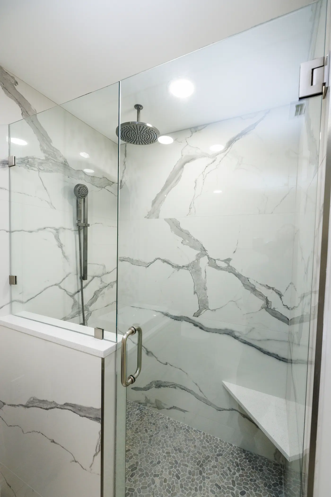 Walk-in shower with large slab marble-look porcelain walls, a frameless door on a half wall with a white quartz cap, a ceiling rain head, a handheld on a slide bar, a floating quartz corner bench, and a gray pebble floor