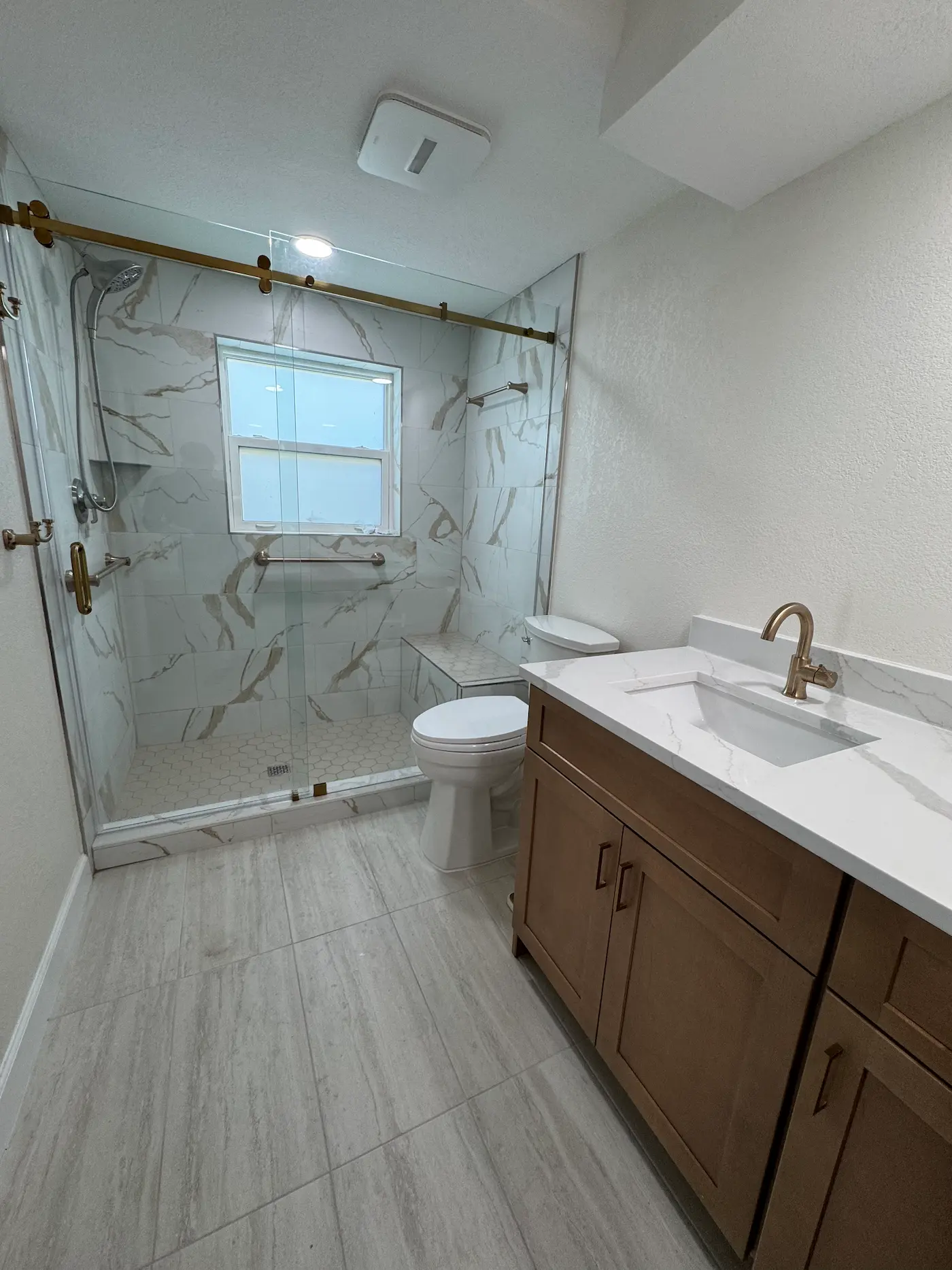 Walk-in shower with a sliding glass door on a brushed brass rail, white and gold marble-look tile, a window, a horizontal grab bar, a tiled corner bench, and a white hexagon floor