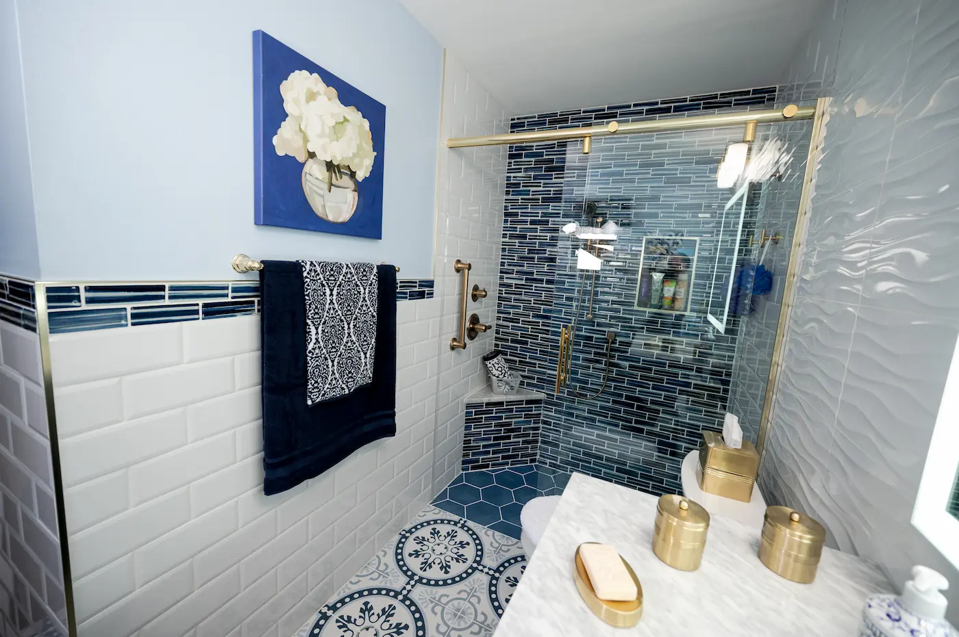 Small bathroom with a walk-in shower in blue glass mosaic tile, a brass frameless sliding door, a corner bench and grab bar, white beveled subway wainscot, and a patterned blue and white floor