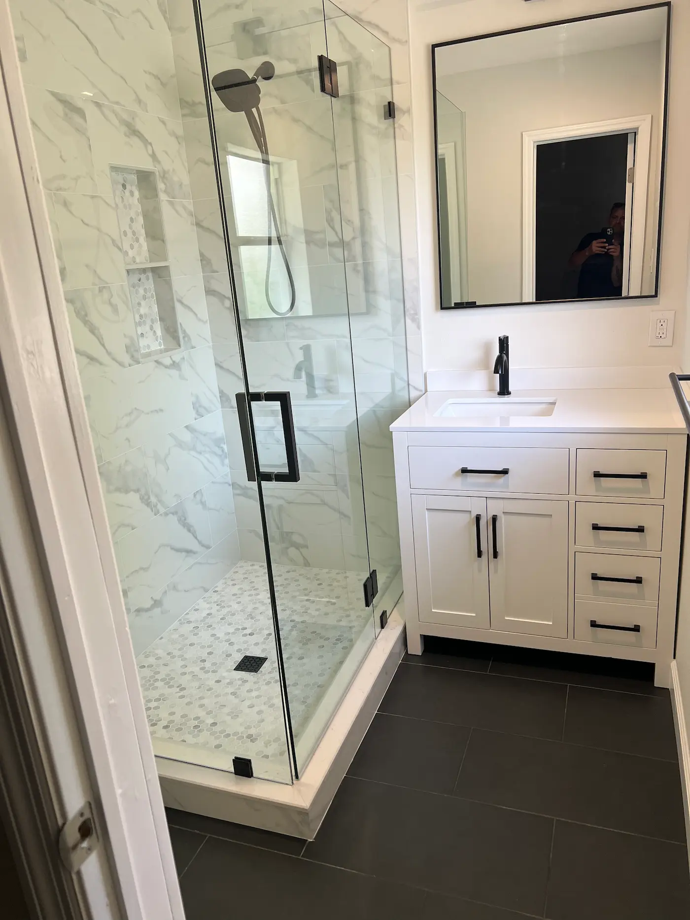 Small corner walk-in shower with frameless glass and matte black hardware, white marble-look porcelain walls, a stacked two-shelf niche lined with mosaic, and a hexagon marble floor beside a white vanity