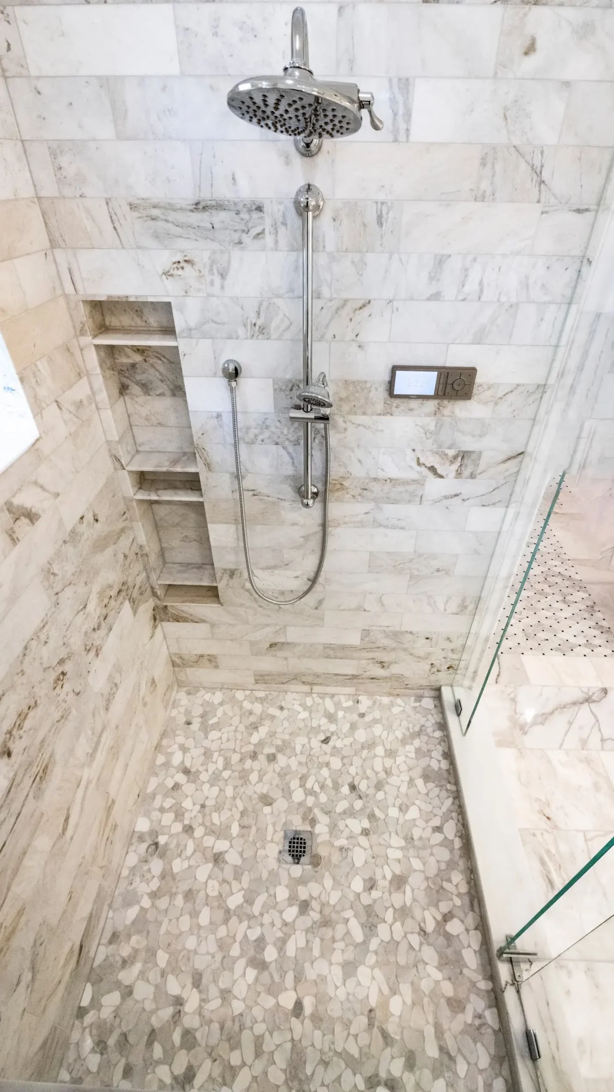 Walk-in shower seen from above with marble-look tile walls, a tall recessed niche with several shelves, a digital shower control panel, a polished nickel rain head and slide bar handheld, and a light pebble floor