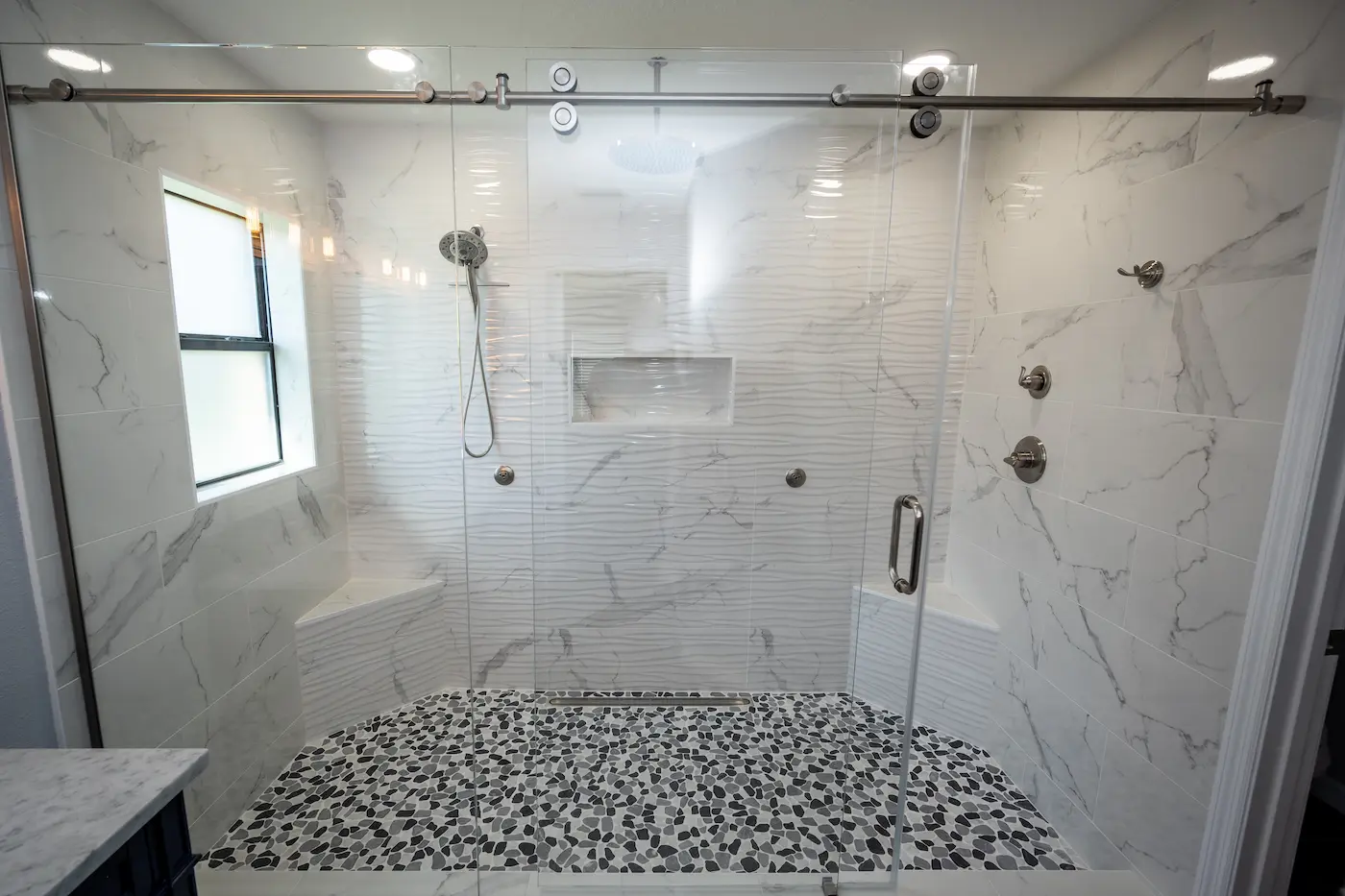Large walk-in shower with a frameless sliding glass door, two corner benches, a wave-textured white accent wall, body sprays, a rain head and handheld, a linear drain, and a black and white pebble floor