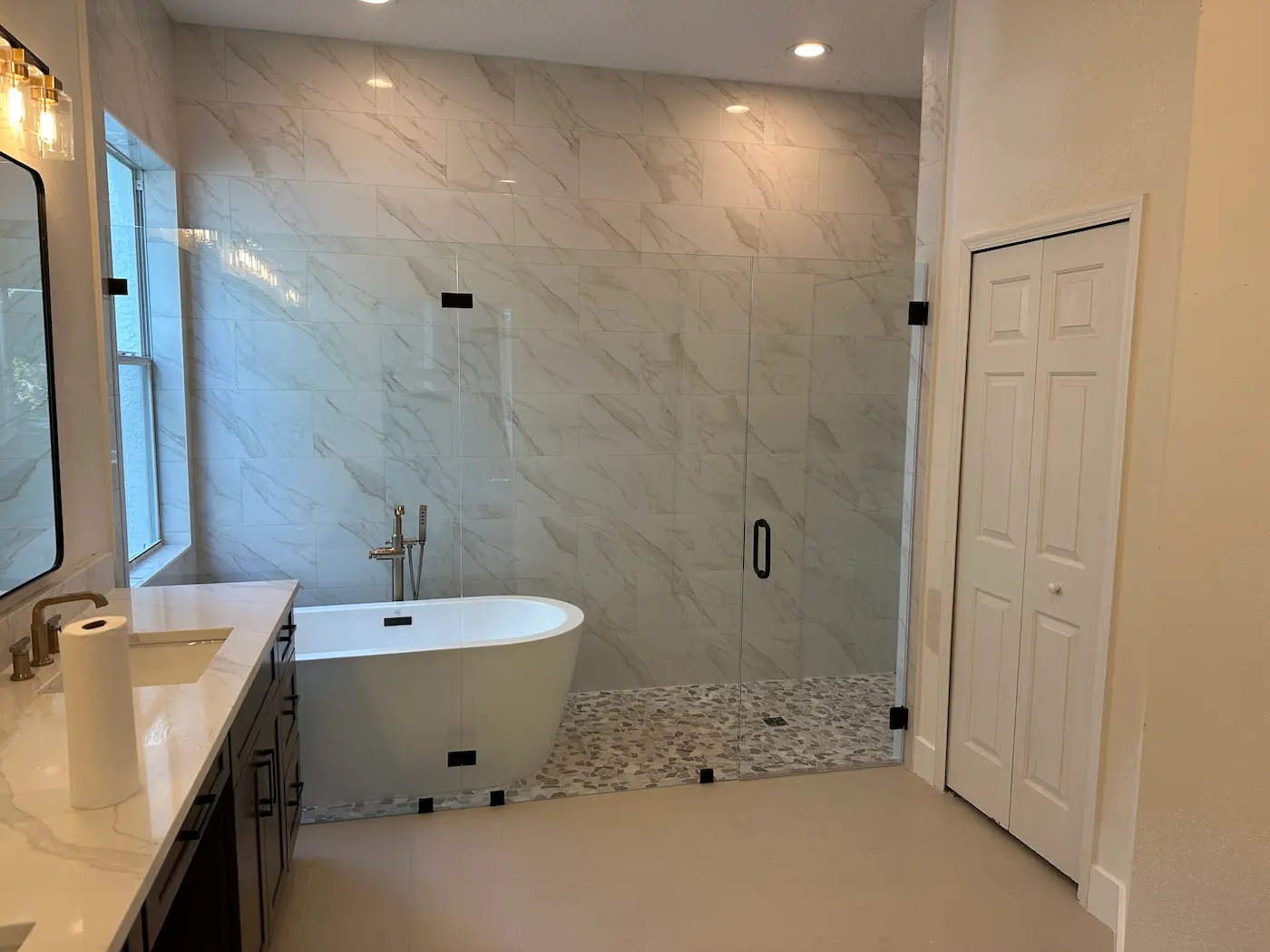 Wet room style walk-in shower with a white freestanding tub and floor-mounted filler inside a frameless glass enclosure, white marble-look wall tile, and a pebble mosaic floor