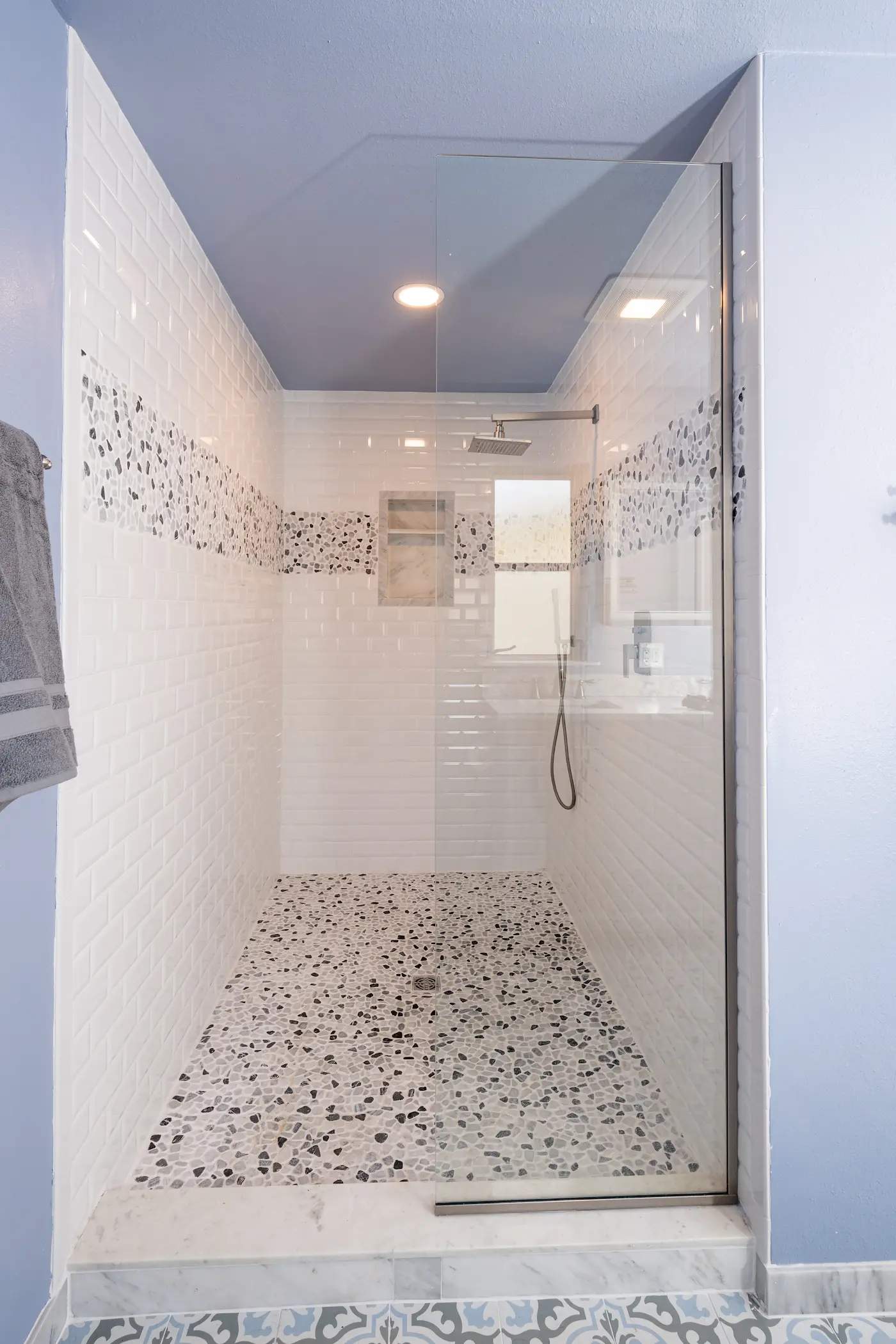 Walk-in shower with white beveled subway tile, a pebble mosaic accent band around all three walls, a matching pebble floor, a single fixed glass panel on a marble curb, a niche, and a window